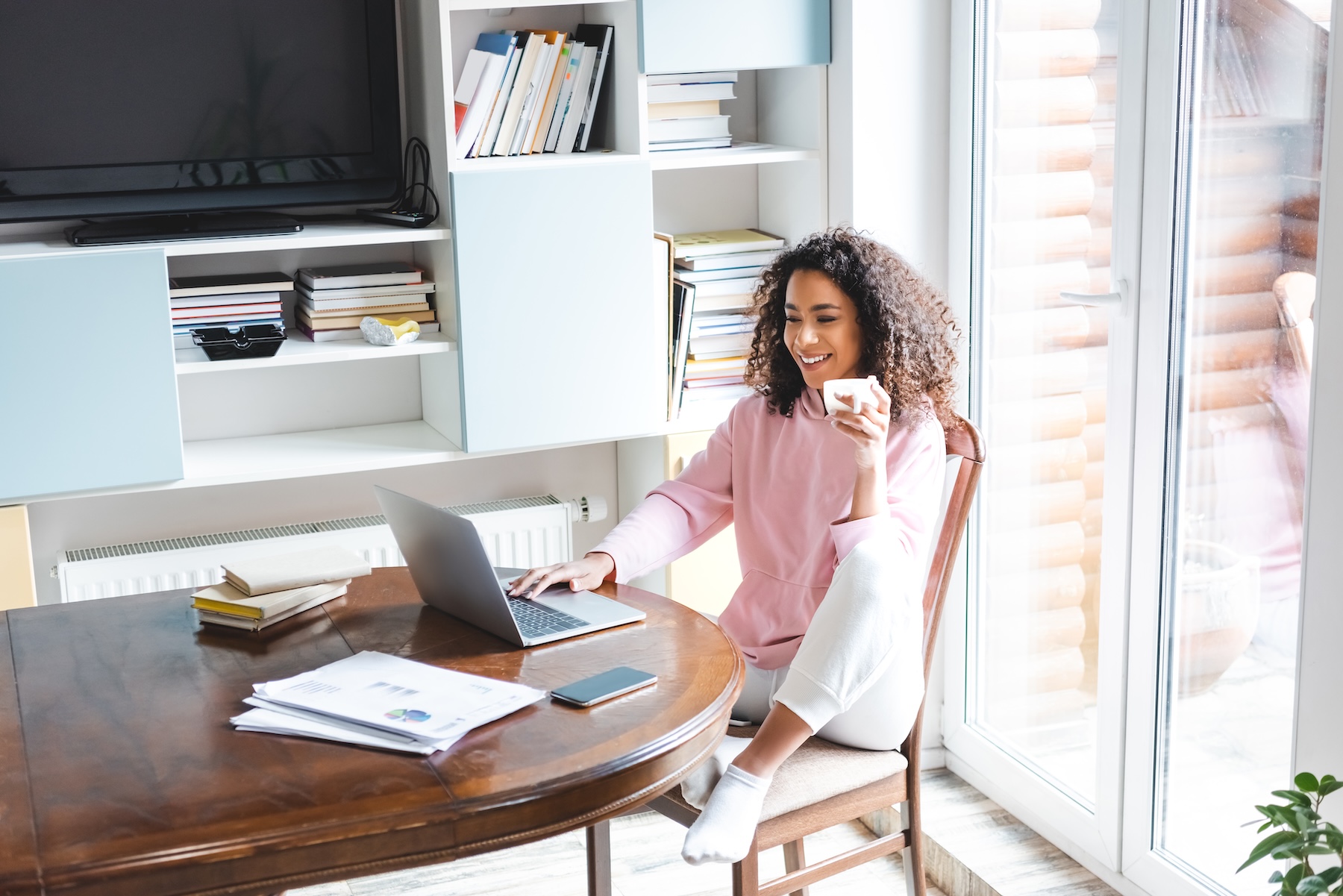Happy African American woman holding cup while using laptop at home