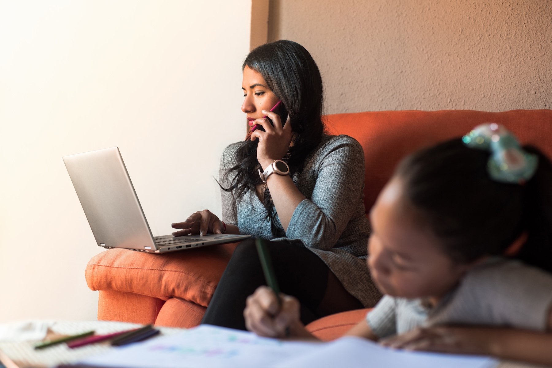 Young mom working at home on laptop, using mobile phone while her daughter does the homework.