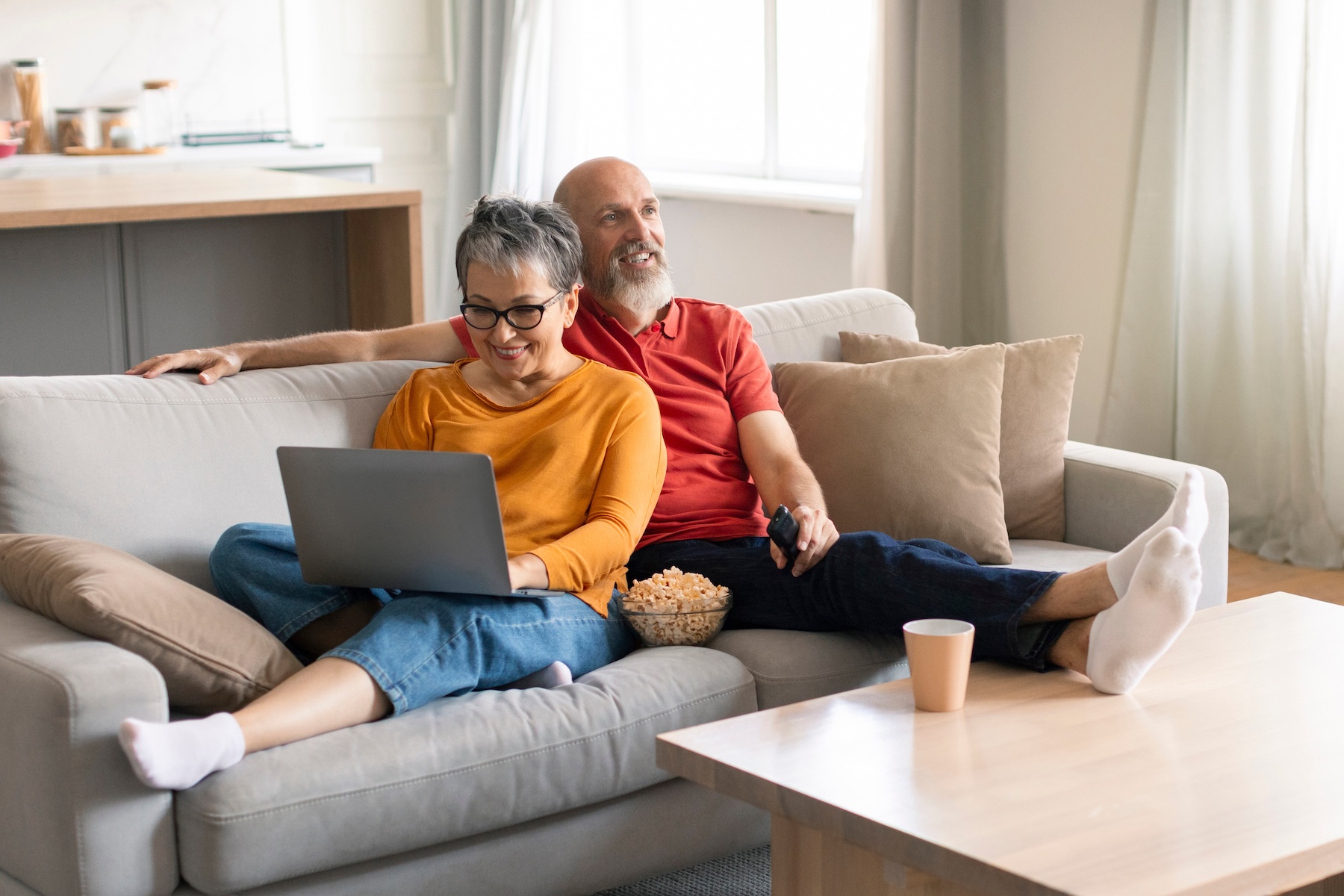 Senior couple resting on couch, one on laptop while the other watches TV