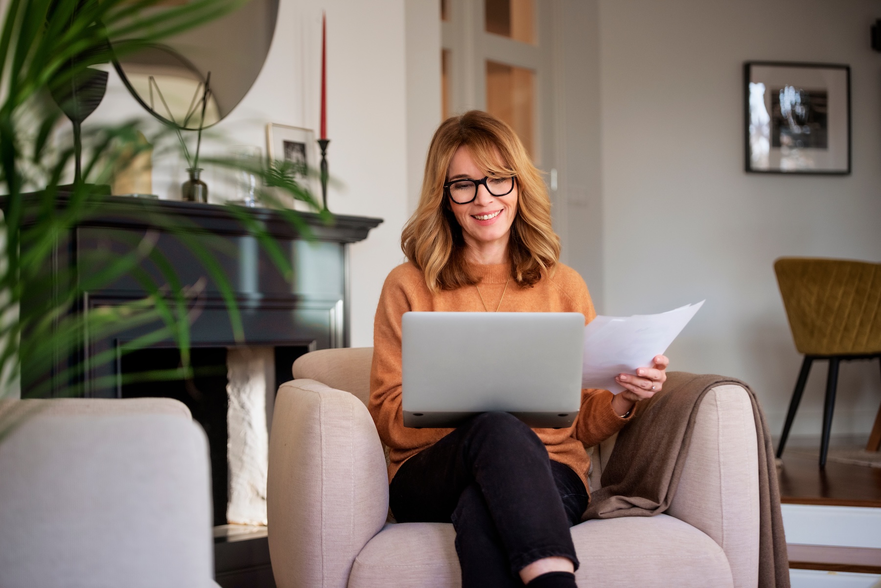 Middle aged woman using laptop and holding document in her hand while sitting at home and working.